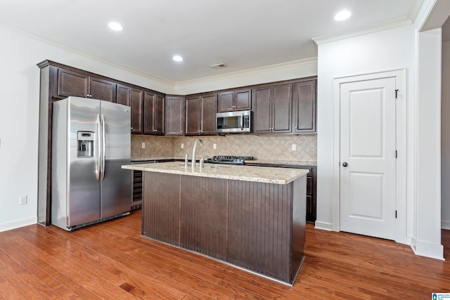 kitchen featuring tasteful backsplash, stainless steel appliances, light stone countertops, dark brown cabinets, and a center island with sink