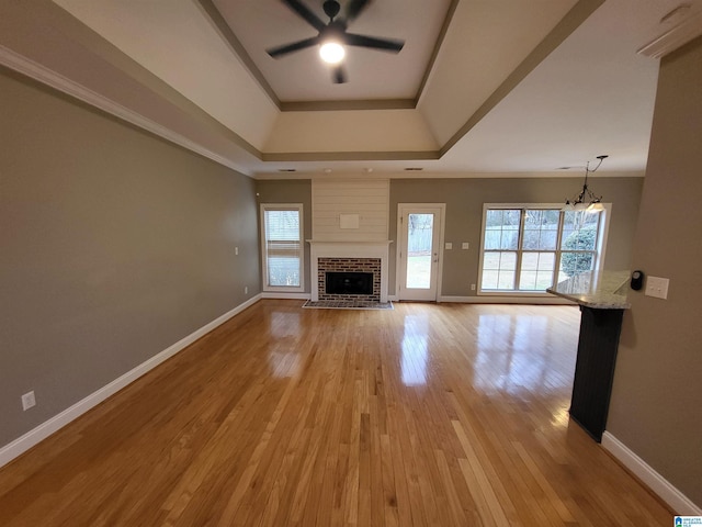 unfurnished living room featuring a raised ceiling, ceiling fan with notable chandelier, a brick fireplace, and light hardwood / wood-style flooring