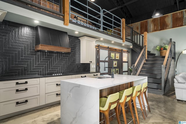 kitchen featuring sink, premium range hood, white cabinets, a center island with sink, and a barn door