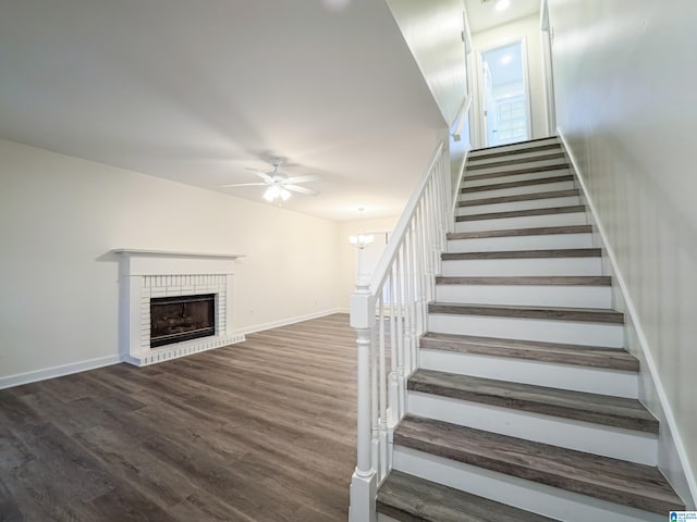 stairway featuring wood-type flooring, ceiling fan with notable chandelier, and a brick fireplace