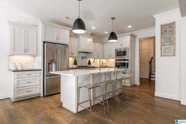 kitchen featuring premium range hood, white cabinetry, stainless steel appliances, a center island with sink, and decorative light fixtures