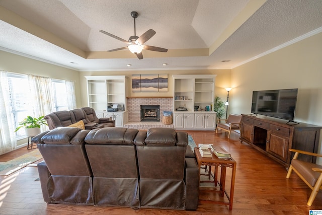 living room featuring hardwood / wood-style flooring, a tray ceiling, crown molding, a brick fireplace, and a textured ceiling