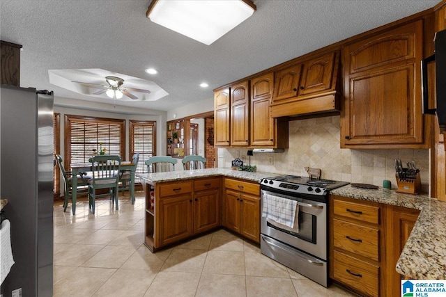 kitchen with light tile patterned flooring, appliances with stainless steel finishes, a tray ceiling, kitchen peninsula, and light stone countertops