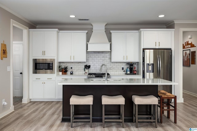 kitchen featuring a kitchen island with sink, stainless steel appliances, white cabinets, and premium range hood