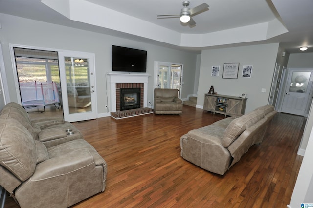 living room with dark wood-type flooring, a fireplace, a tray ceiling, and ceiling fan