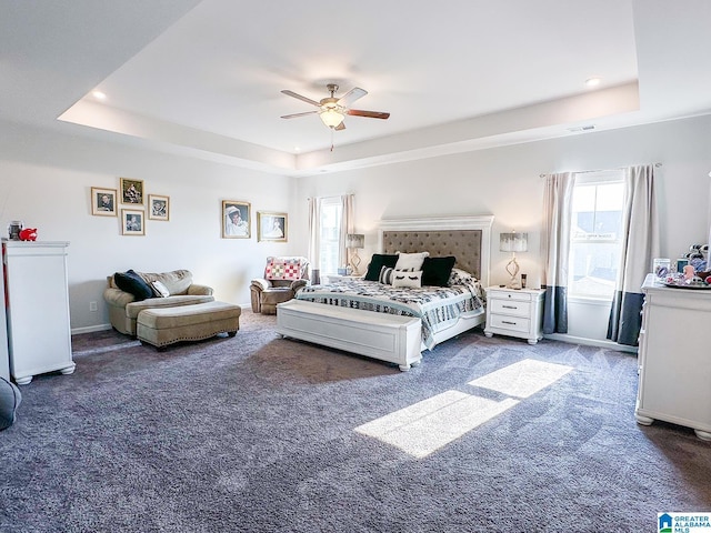 bedroom featuring a raised ceiling, ceiling fan, dark carpet, and multiple windows