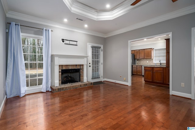 unfurnished living room featuring dark wood-type flooring, a brick fireplace, ornamental molding, a tray ceiling, and ceiling fan