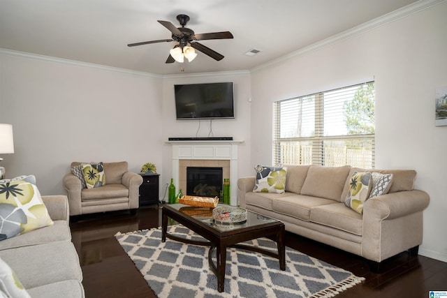 living room with crown molding, ceiling fan, a tiled fireplace, and dark wood-type flooring