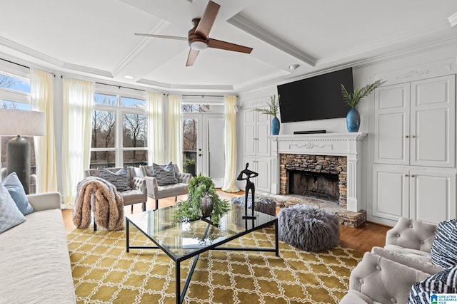 living room featuring a stone fireplace, beamed ceiling, wood-type flooring, ornamental molding, and ceiling fan