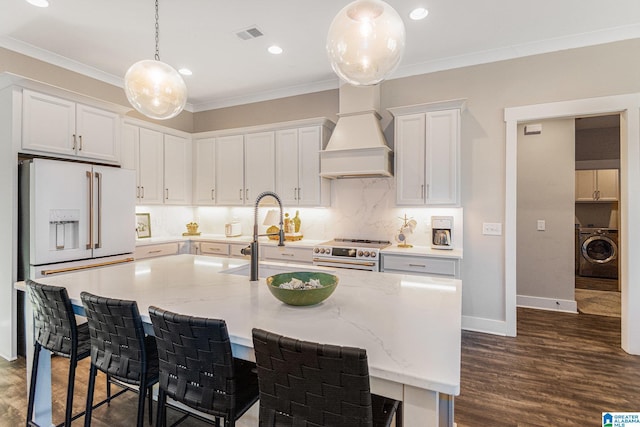 kitchen with custom exhaust hood, washer / clothes dryer, a kitchen island with sink, white cabinets, and high quality appliances