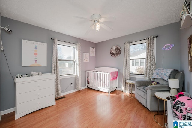 bedroom featuring a nursery area, ceiling fan, multiple windows, and light hardwood / wood-style flooring