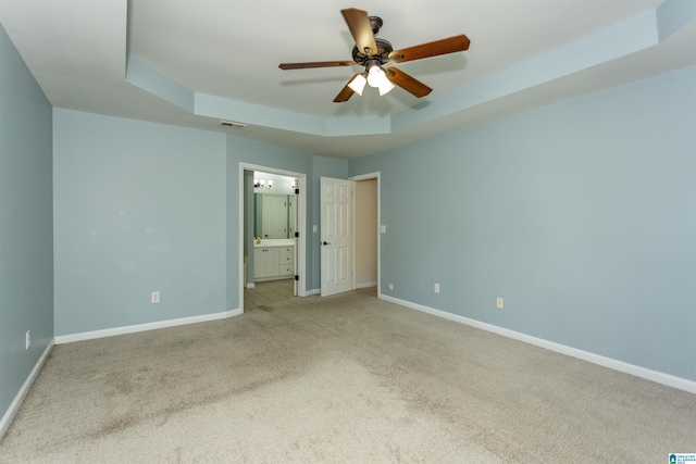 unfurnished bedroom featuring ceiling fan, ensuite bathroom, a tray ceiling, and light carpet