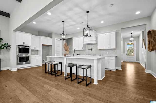 kitchen with pendant lighting, stainless steel double oven, a kitchen island with sink, and white cabinets