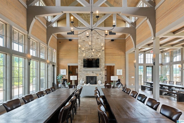 dining space with wooden walls, a towering ceiling, a notable chandelier, a stone fireplace, and beam ceiling