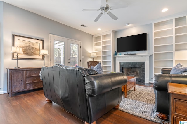 living room with built in shelves, a tiled fireplace, ceiling fan, dark wood-type flooring, and french doors