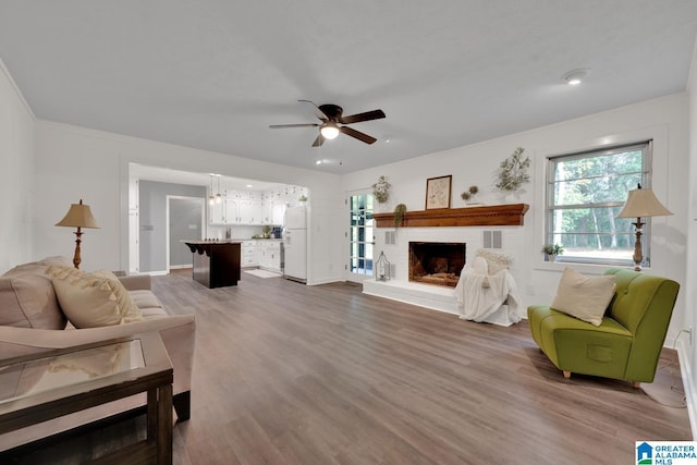 living room featuring a brick fireplace, wood-type flooring, ornamental molding, and ceiling fan