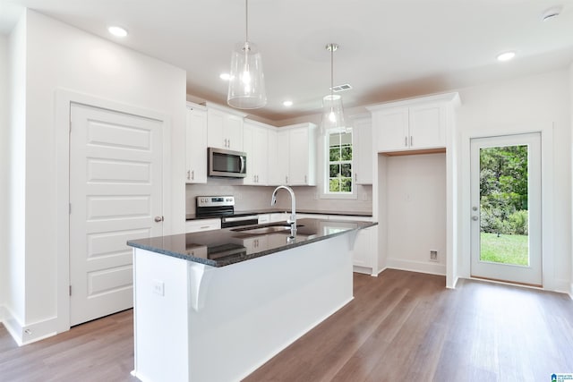 kitchen featuring a center island with sink, appliances with stainless steel finishes, white cabinetry, dark stone counters, and decorative light fixtures