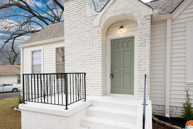 doorway to property featuring a shingled roof, a chimney, and brick siding