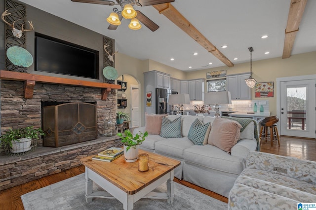 living area with arched walkways, wood finished floors, a stone fireplace, beam ceiling, and recessed lighting