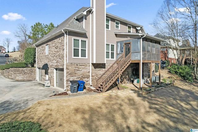 back of house with concrete driveway, a sunroom, a chimney, stairs, and brick siding