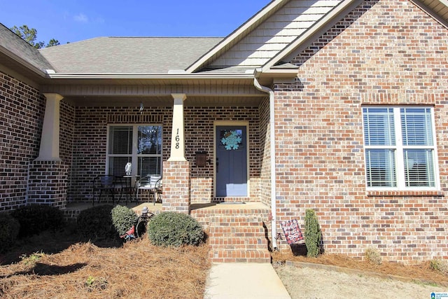 doorway to property with covered porch, a shingled roof, and brick siding