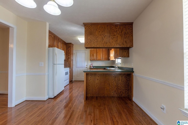 kitchen with a peninsula, freestanding refrigerator, brown cabinetry, dark countertops, and dark wood finished floors