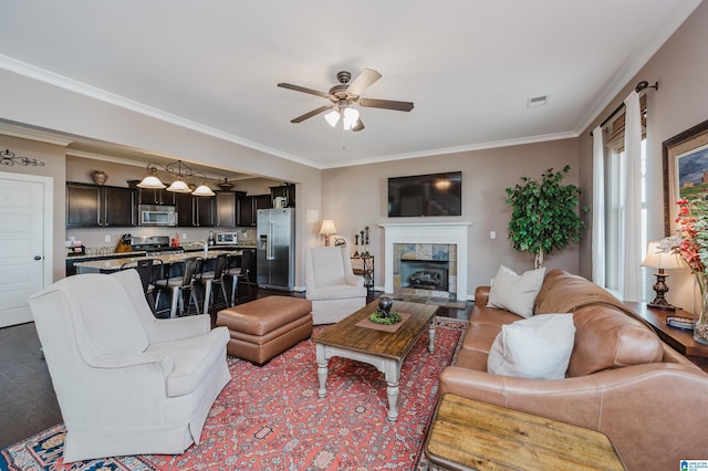 living room with ceiling fan, wood finished floors, visible vents, ornamental molding, and a tiled fireplace