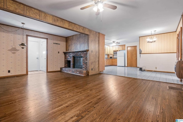 unfurnished living room featuring ceiling fan with notable chandelier, a brick fireplace, wood finished floors, and wallpapered walls