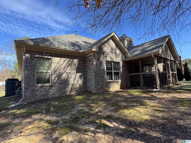 back of property featuring a shingled roof, central AC unit, a sunroom, a chimney, and brick siding