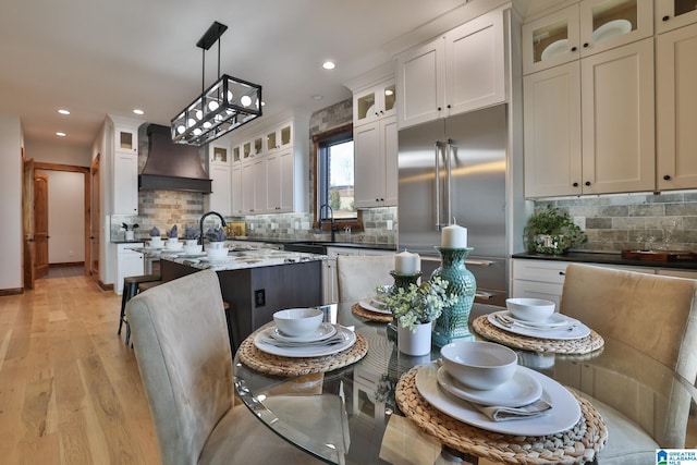 kitchen featuring hanging light fixtures, custom exhaust hood, stainless steel built in refrigerator, glass insert cabinets, and an island with sink