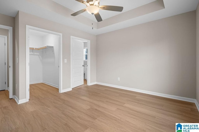 unfurnished bedroom with baseboards, a tray ceiling, a walk in closet, light wood-style floors, and a closet