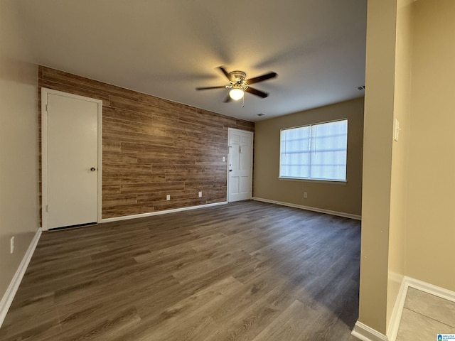 unfurnished room with an accent wall, dark wood-type flooring, wood walls, a ceiling fan, and baseboards