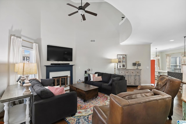 living area with ceiling fan, a high ceiling, wood finished floors, visible vents, and a glass covered fireplace