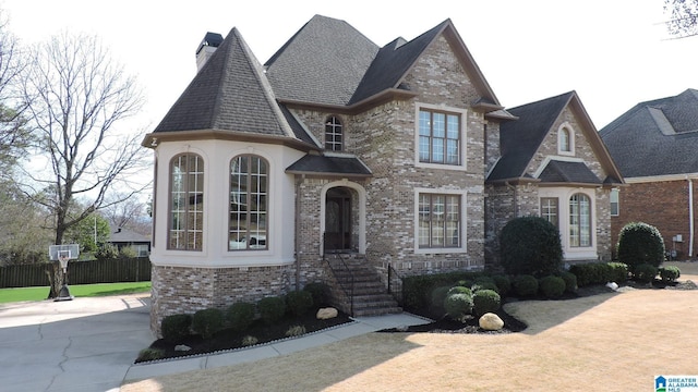 french country home featuring roof with shingles, brick siding, a chimney, and fence