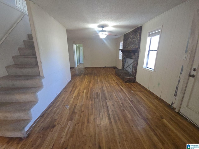 unfurnished living room with a textured ceiling, dark wood-style flooring, a ceiling fan, stairs, and a brick fireplace