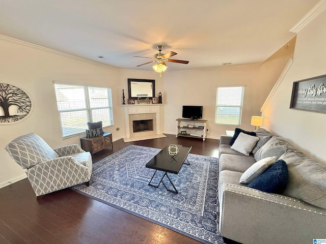 living area with dark wood finished floors, crown molding, a tiled fireplace, a ceiling fan, and baseboards