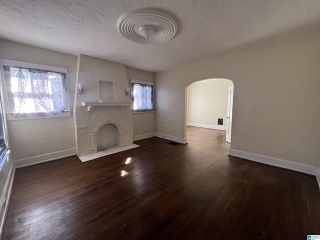 unfurnished living room featuring dark wood-style floors, arched walkways, a brick fireplace, a textured ceiling, and baseboards