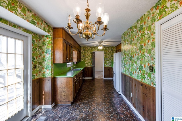 kitchen with brown cabinetry, a wainscoted wall, a sink, and wallpapered walls