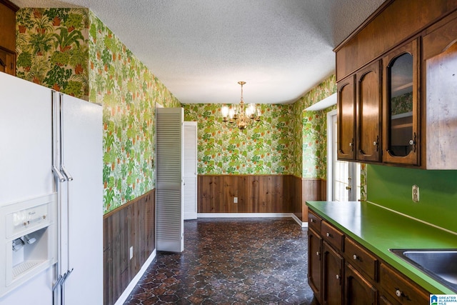 kitchen with a textured ceiling, a notable chandelier, white refrigerator with ice dispenser, wainscoting, and wallpapered walls