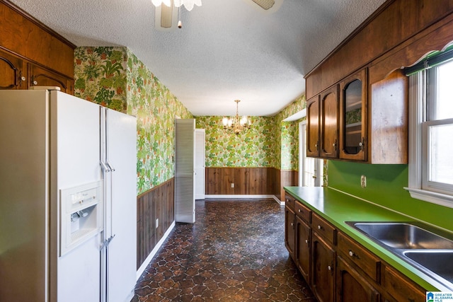 kitchen featuring a textured ceiling, white refrigerator with ice dispenser, a sink, wainscoting, and wallpapered walls