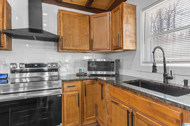 kitchen featuring stainless steel appliances, brown cabinetry, a sink, wall chimney range hood, and dark stone countertops