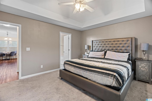 bedroom featuring light carpet, baseboards, ensuite bathroom, a tray ceiling, and ceiling fan with notable chandelier