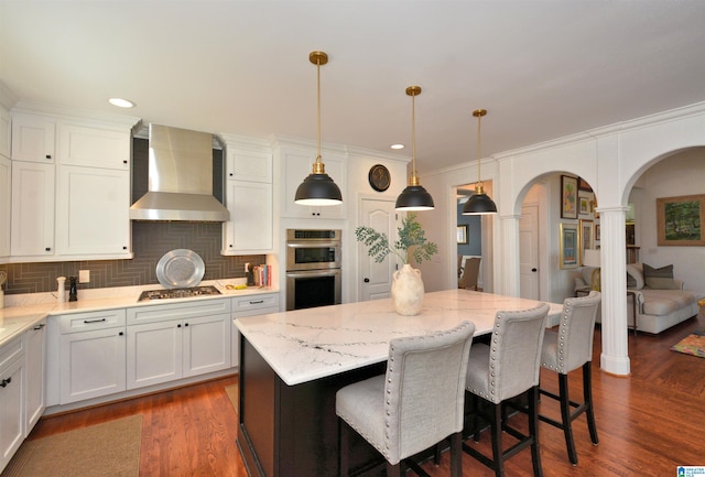 kitchen featuring arched walkways, white cabinets, wall chimney exhaust hood, a kitchen island, and appliances with stainless steel finishes