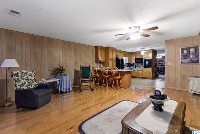 living room featuring ceiling fan, light wood finished floors, visible vents, and wooden walls