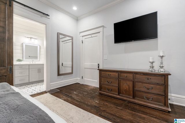 bedroom with a barn door, dark wood-type flooring, ornamental molding, ensuite bath, and baseboards