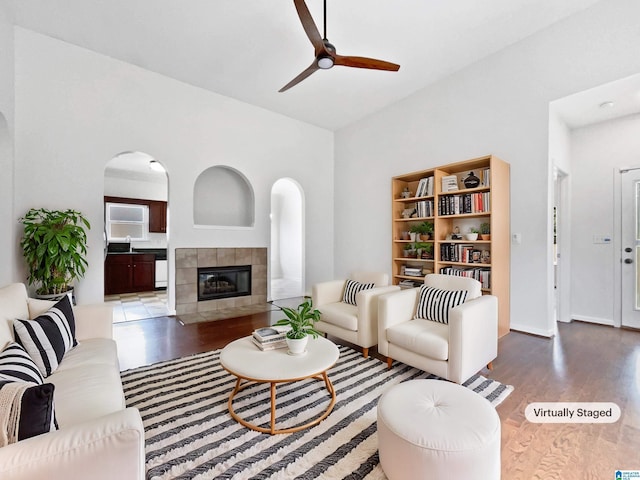 living room with arched walkways, wood finished floors, a ceiling fan, baseboards, and a tiled fireplace