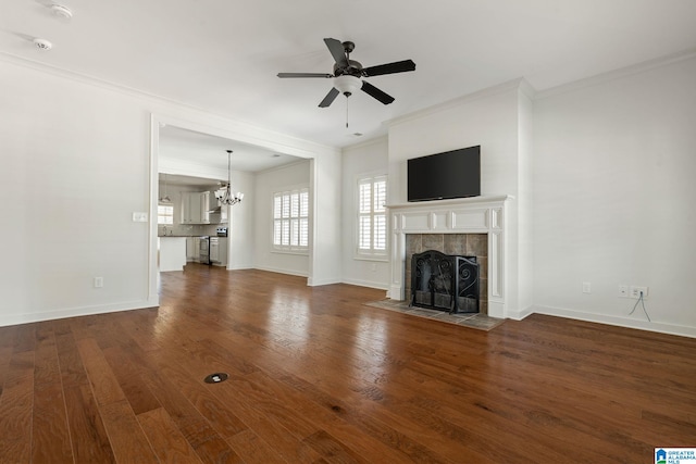 unfurnished living room featuring crown molding, dark wood finished floors, a tiled fireplace, and ceiling fan with notable chandelier