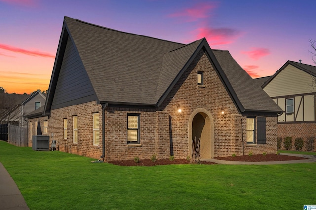 english style home with a yard, a shingled roof, central AC unit, and brick siding