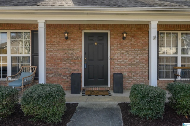 entrance to property featuring covered porch, brick siding, and roof with shingles