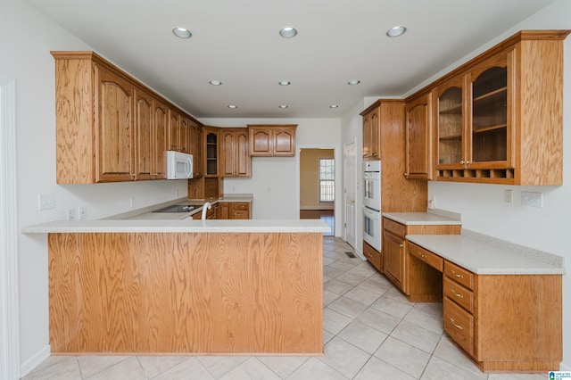 kitchen featuring built in desk, glass insert cabinets, brown cabinetry, white appliances, and a peninsula